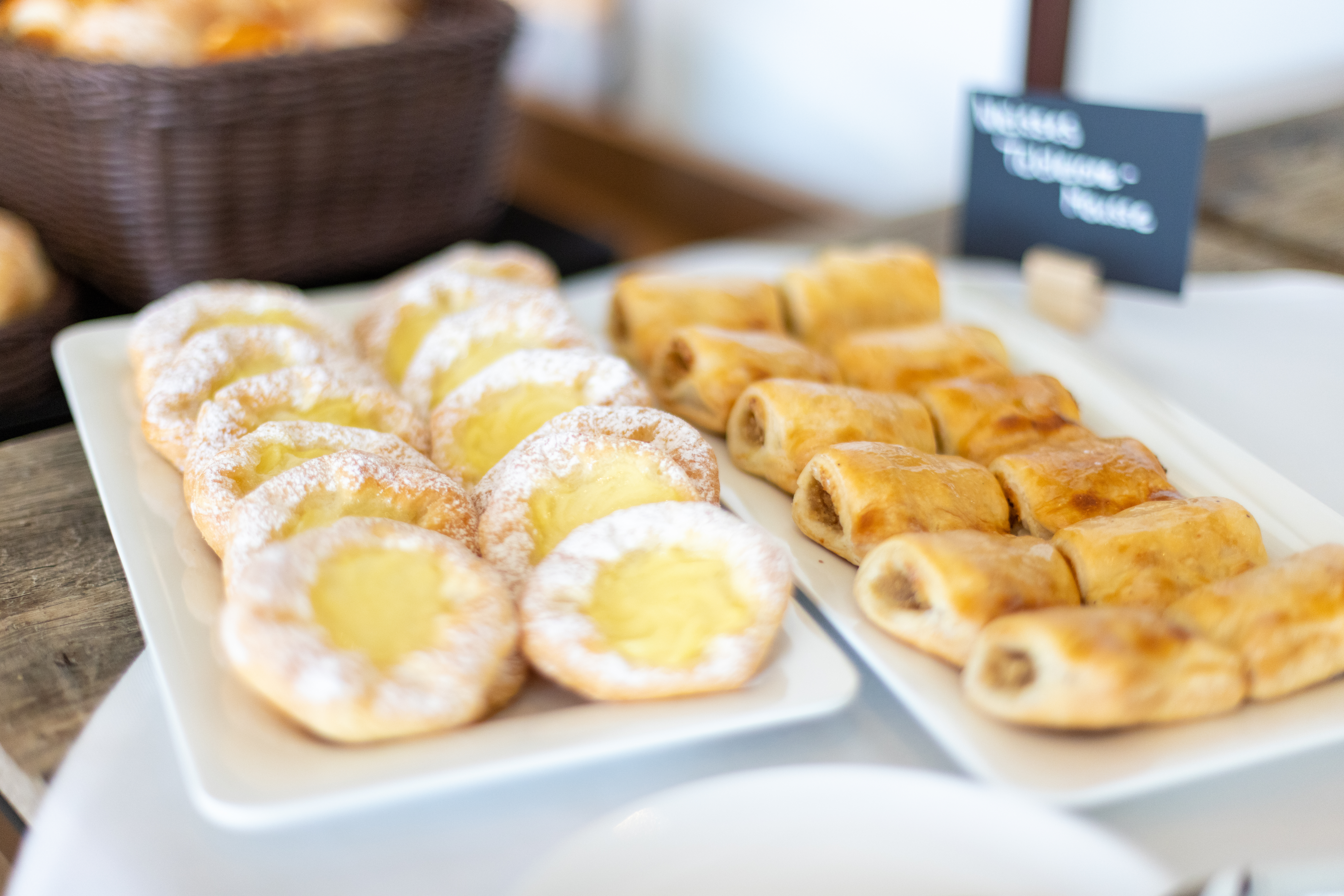 Brunch-Buffet: Zwei Platten mit Gebäck. Eine Platte zeigt runde, puderzuckerbestreute Teilchen mit gelber Füllung. Die andere Platte präsentiert längliche, goldbraune gefüllte Teigrollen. Im Hintergrund ein Brotkorb und eine Tafel.